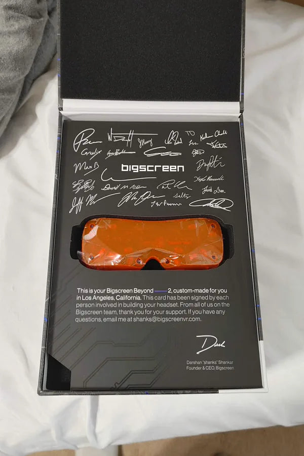 An orange Beyond 2 in its opened box with the signed card, on a bed